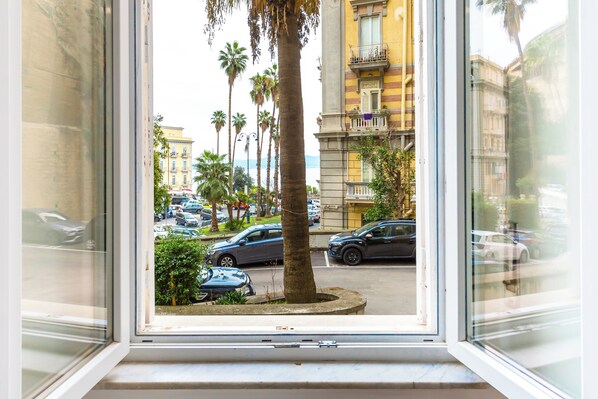 A window view of a street with parked cars, palm trees, and buildings. - San Luigi cozy flat - private parking (Naples)