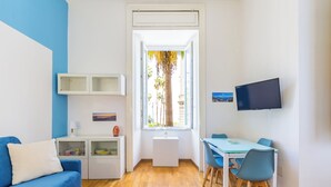 A modern living room with a blue sofa, a dining table with chairs, a TV mounted on the wall, and a window with a view of palm trees. - San Luigi cozy flat - private parking (Naples)