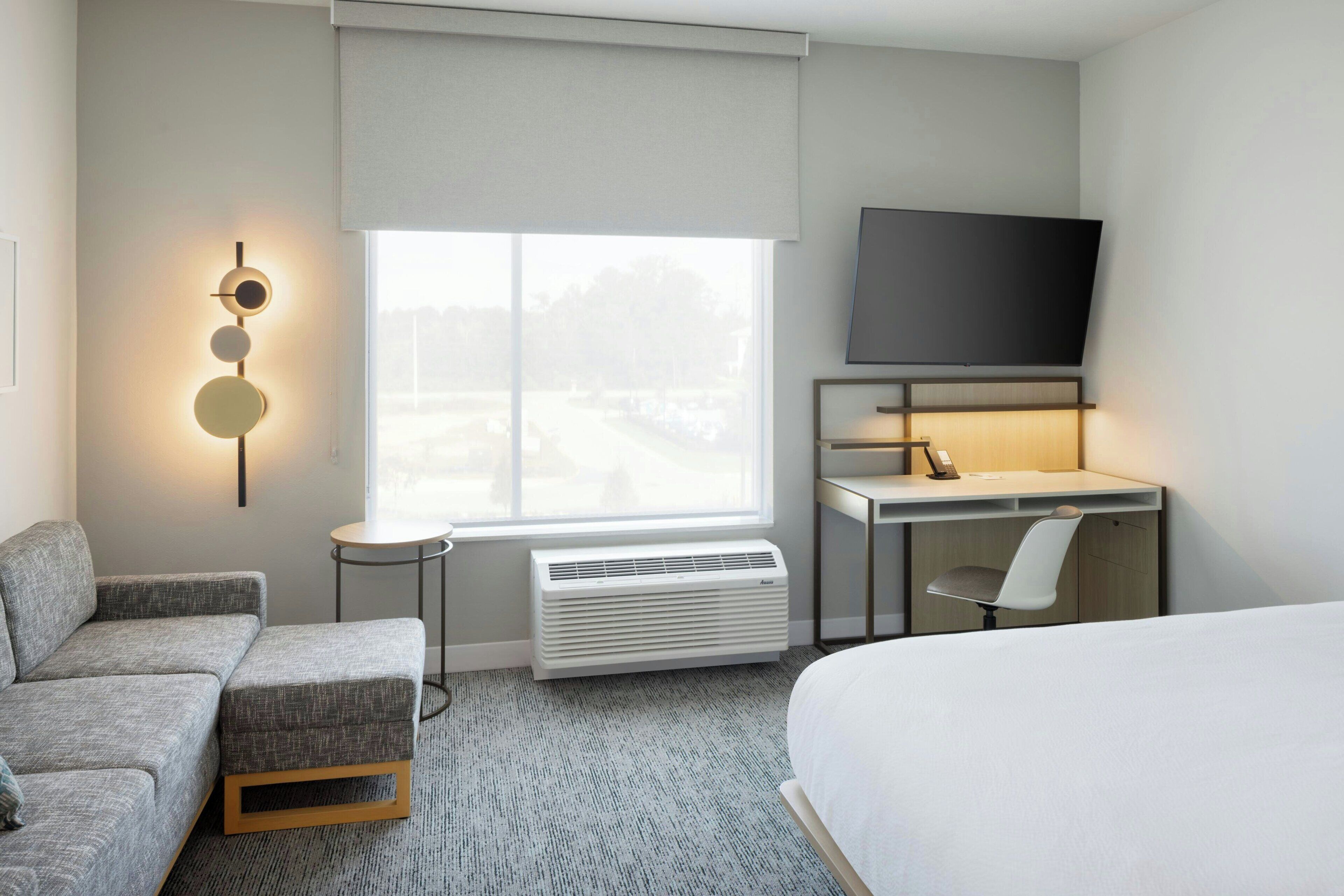 Ett hotellrum med en grå soffa, en säng, ett skrivbord med en stol, en platt-TV och ett fönster med persienner.