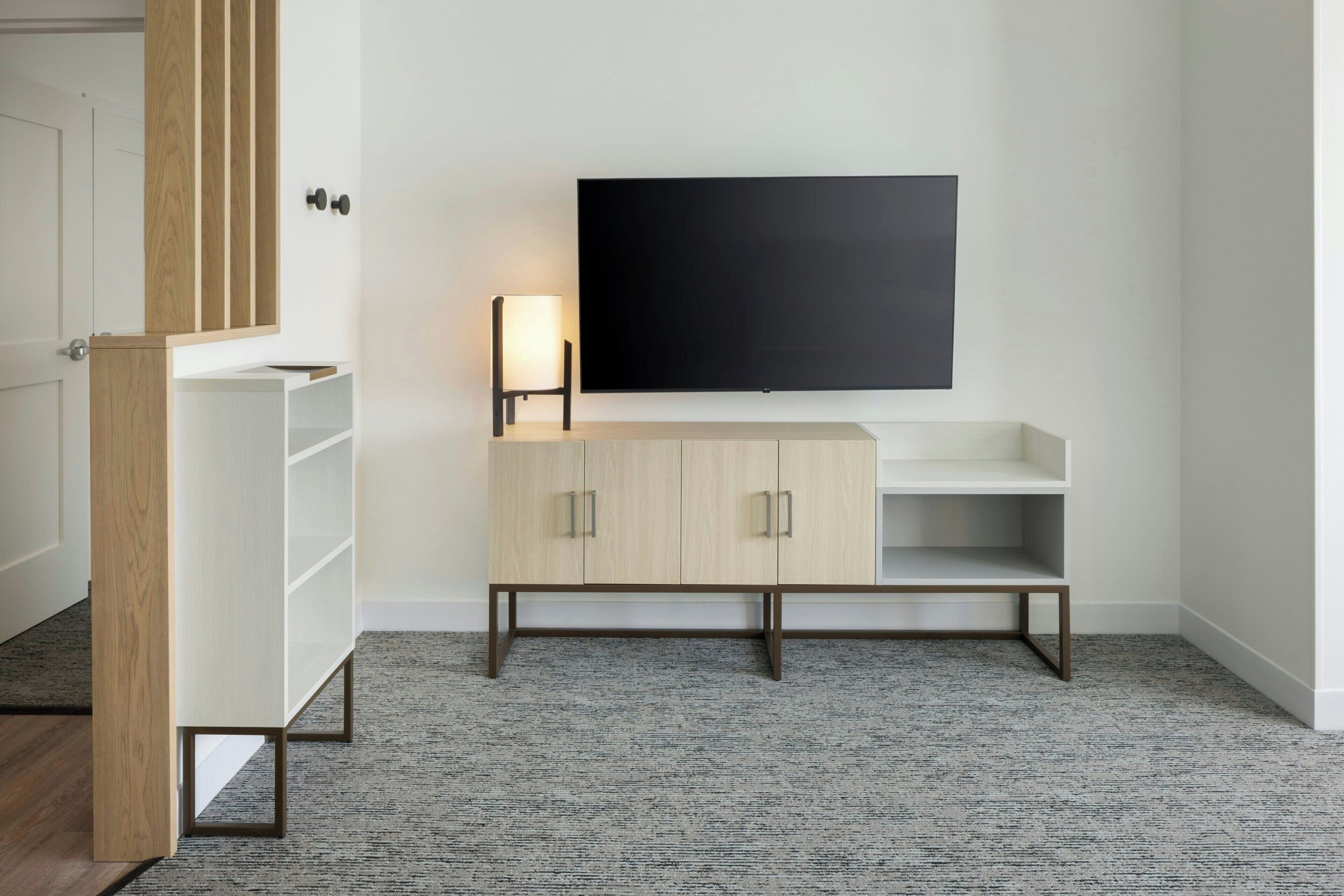 A modern room with a flat-screen TV mounted on the wall, a wooden cabinet with open shelves, and a carpeted floor.