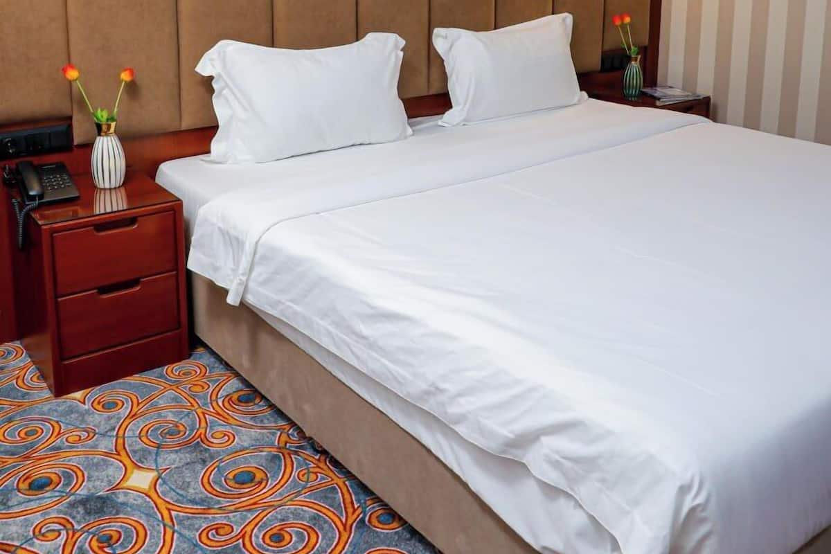 A hotel room with a large bed, two bedside tables, a nightstand, a wall-mounted lamp, and a decorative vase with flowers.