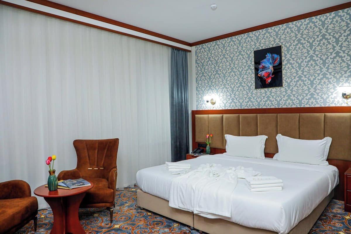 A hotel room with a large bed, a brown armchair, a small table with a vase of flowers, and a decorative wall with a butterfly painting.
