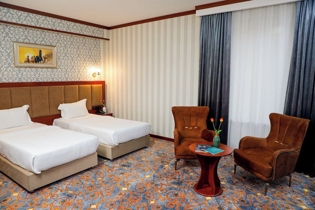 A hotel room with two beds, a brown armchair, a red table, and a patterned carpet.