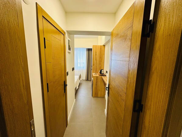 A hallway with wooden doors leading to a room with a bed, a small table, and a window with curtains. - YAKIN PANSİYON (Amasra)