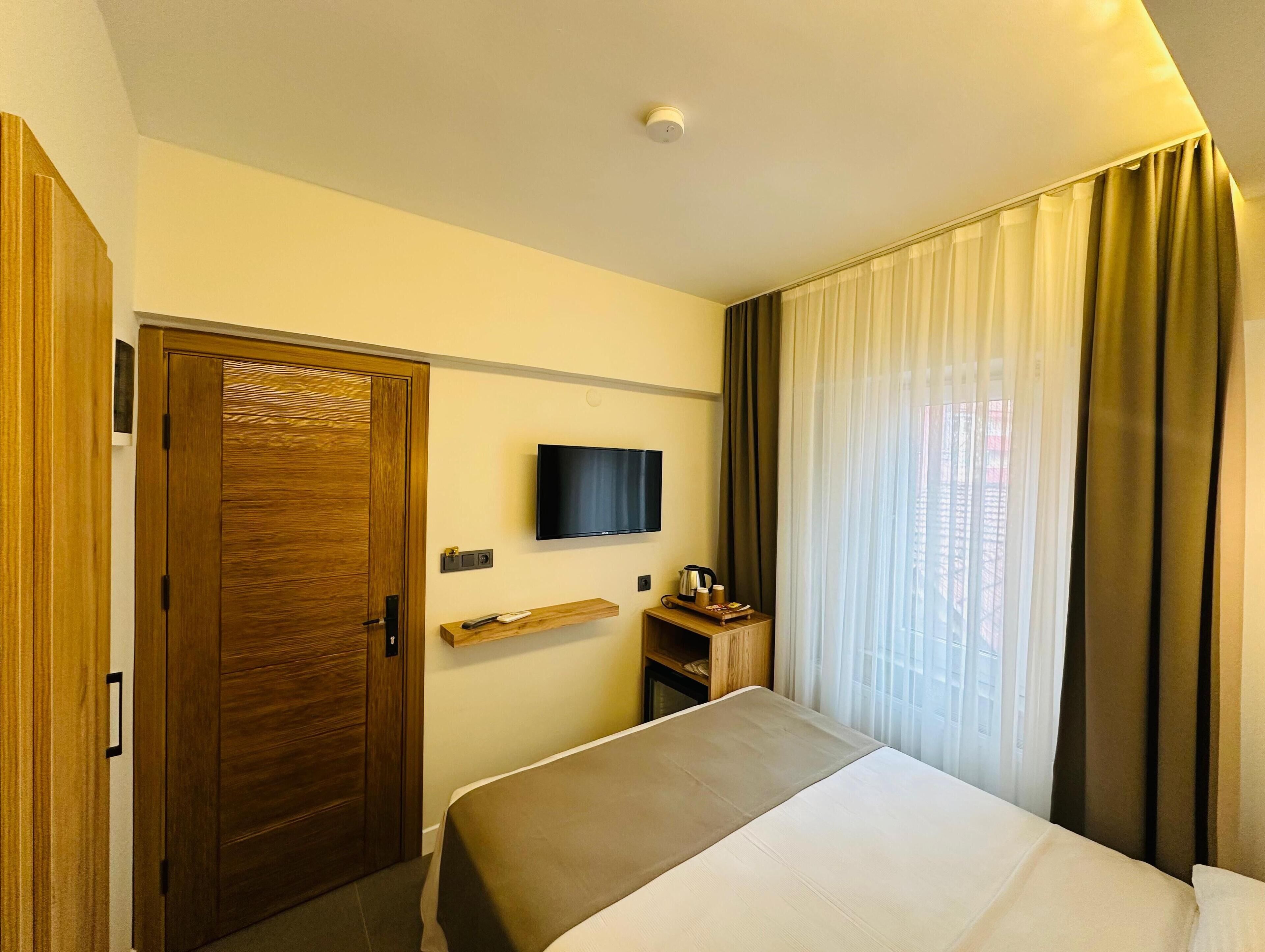 A hotel room with a bed, a television mounted on the wall, a wooden wardrobe, and a window with curtains.