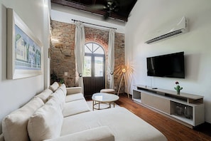 A living room with a stone wall, a large window with curtains, a white sofa, a round coffee table, a TV on a stand, and a cactus on the stand.