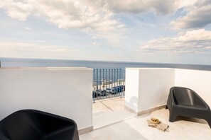 A balcony with a view of the sea, a chair, a small table with a bowl, and a blue railing.