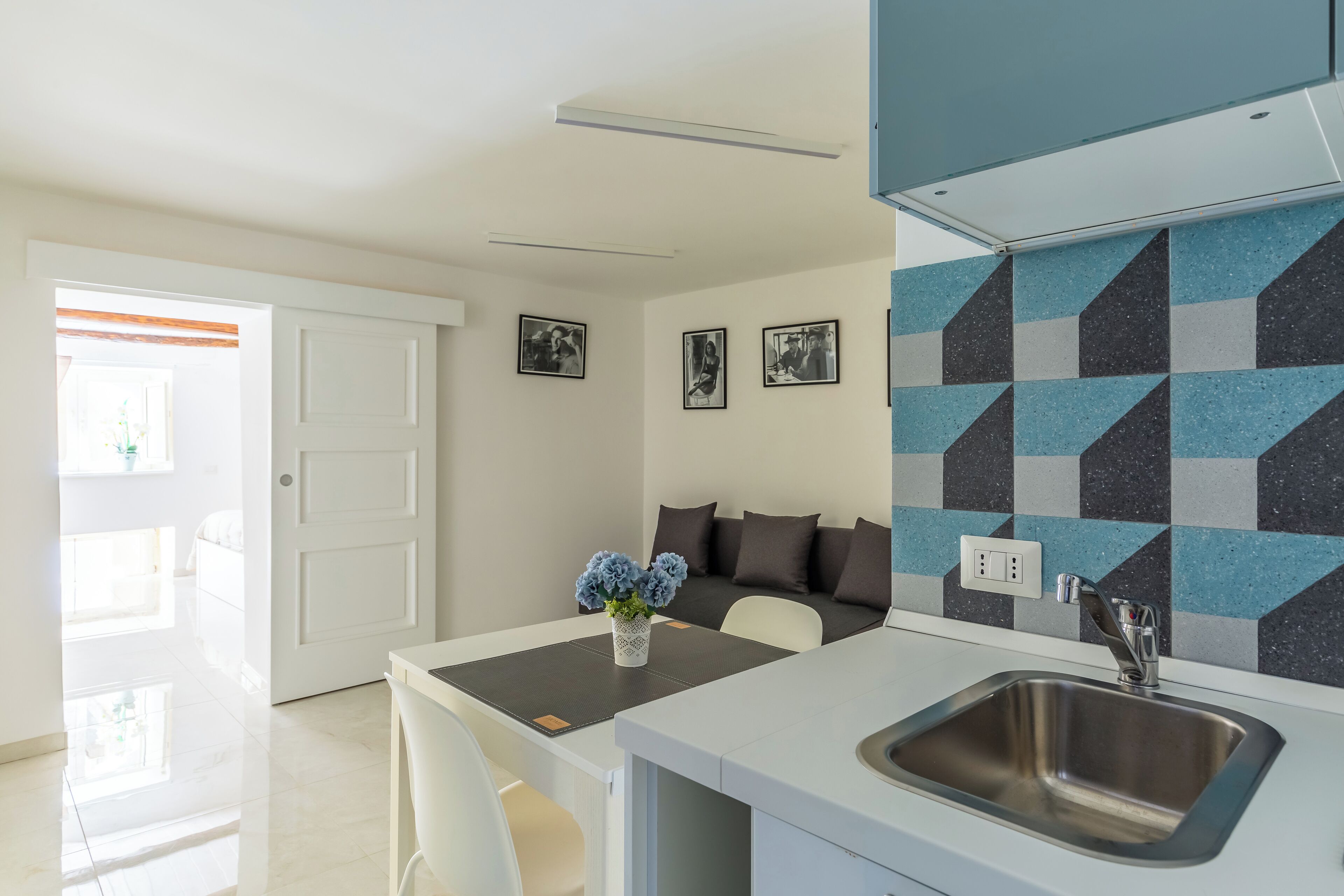 A modern kitchen with a sink, a dining area with a table and chairs, and a living area with a sofa and cushions.