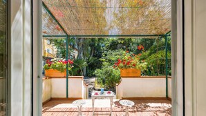 Un patio soleado con una mesa y dos sillas, rodeado de plantas en macetas y vistas a un jardín.