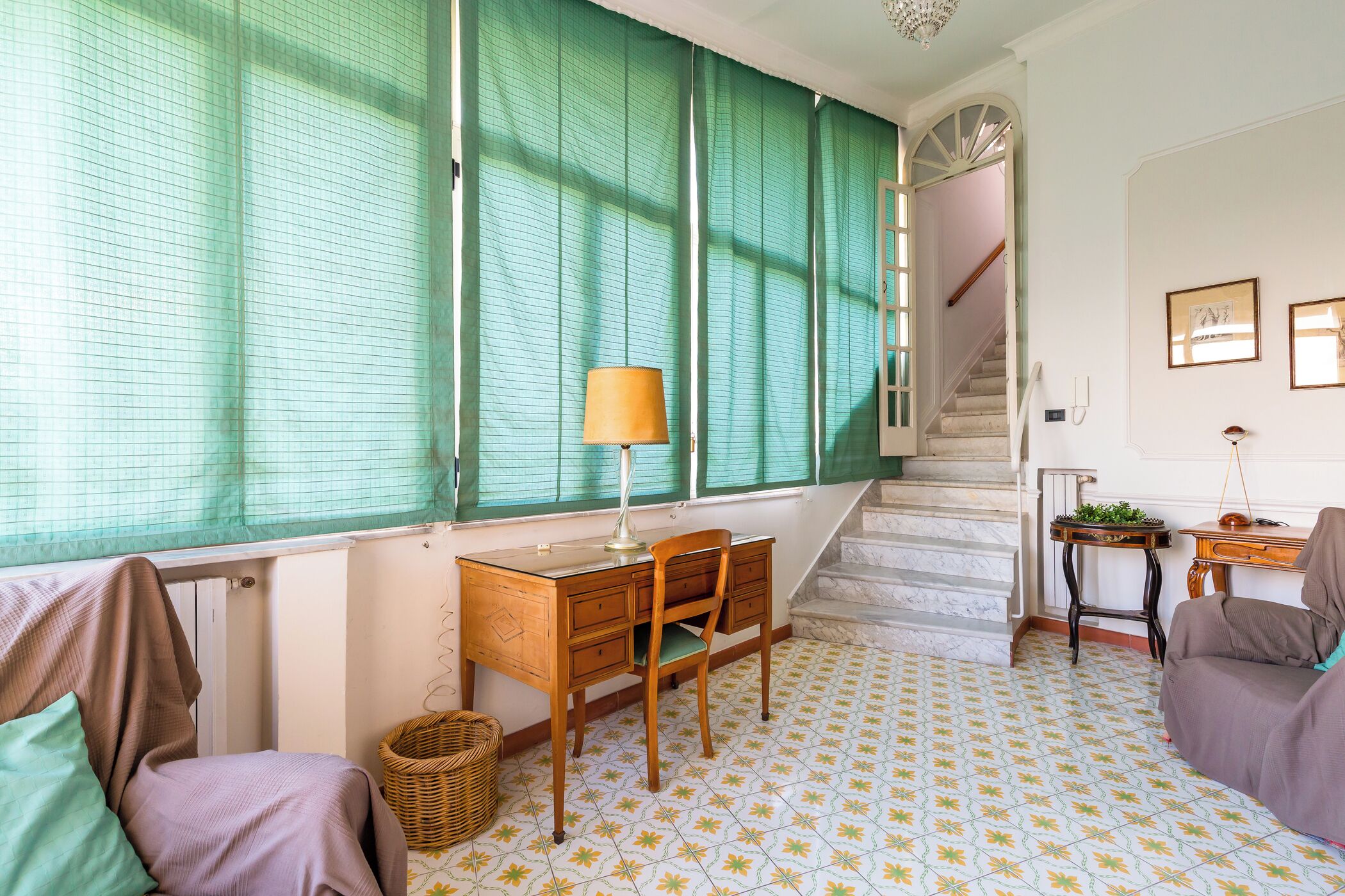 A room with a wooden desk, a chair, a lamp, a wicker basket, and a staircase leading to another level.