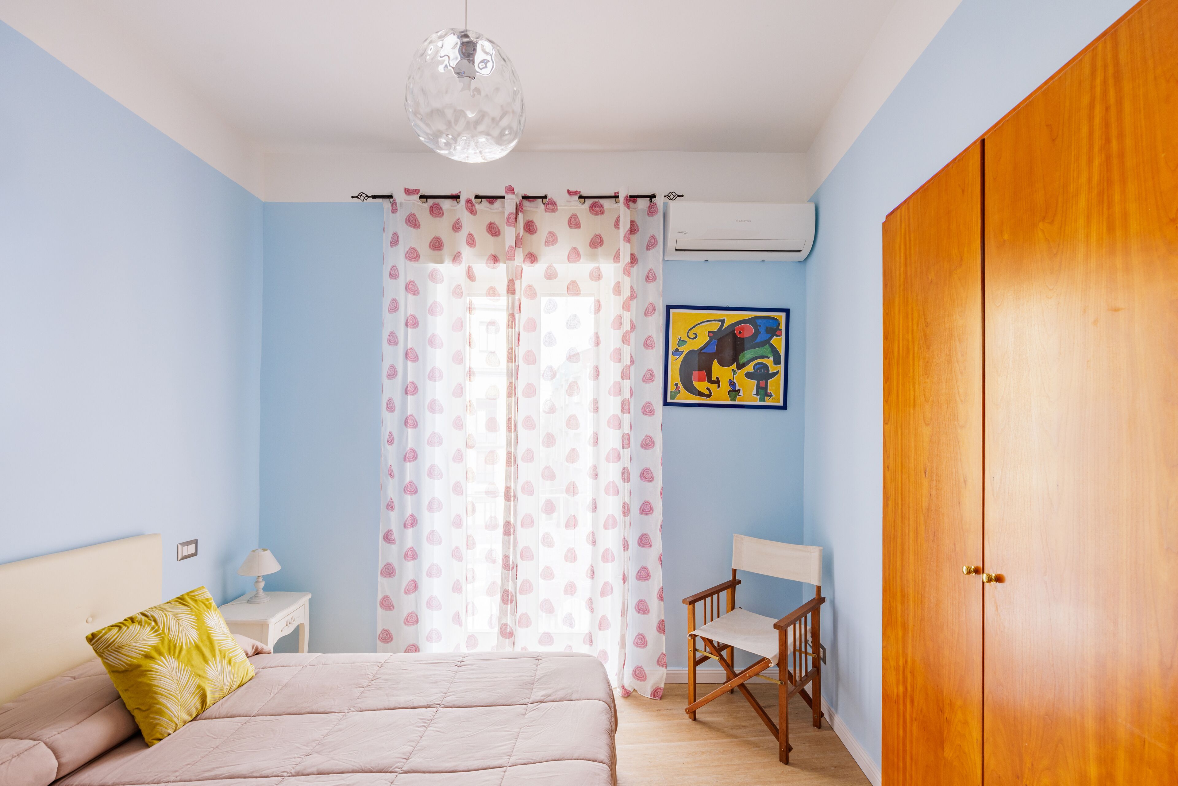 A bedroom with a bed, a chair, a nightstand, a lamp, a wardrobe, and a painting on the wall.