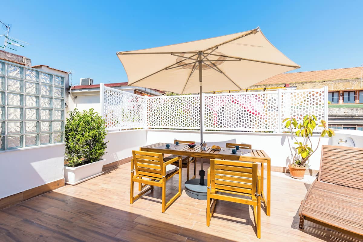 A rooftop terrace with a wooden table and chairs, a large umbrella, and potted plants.