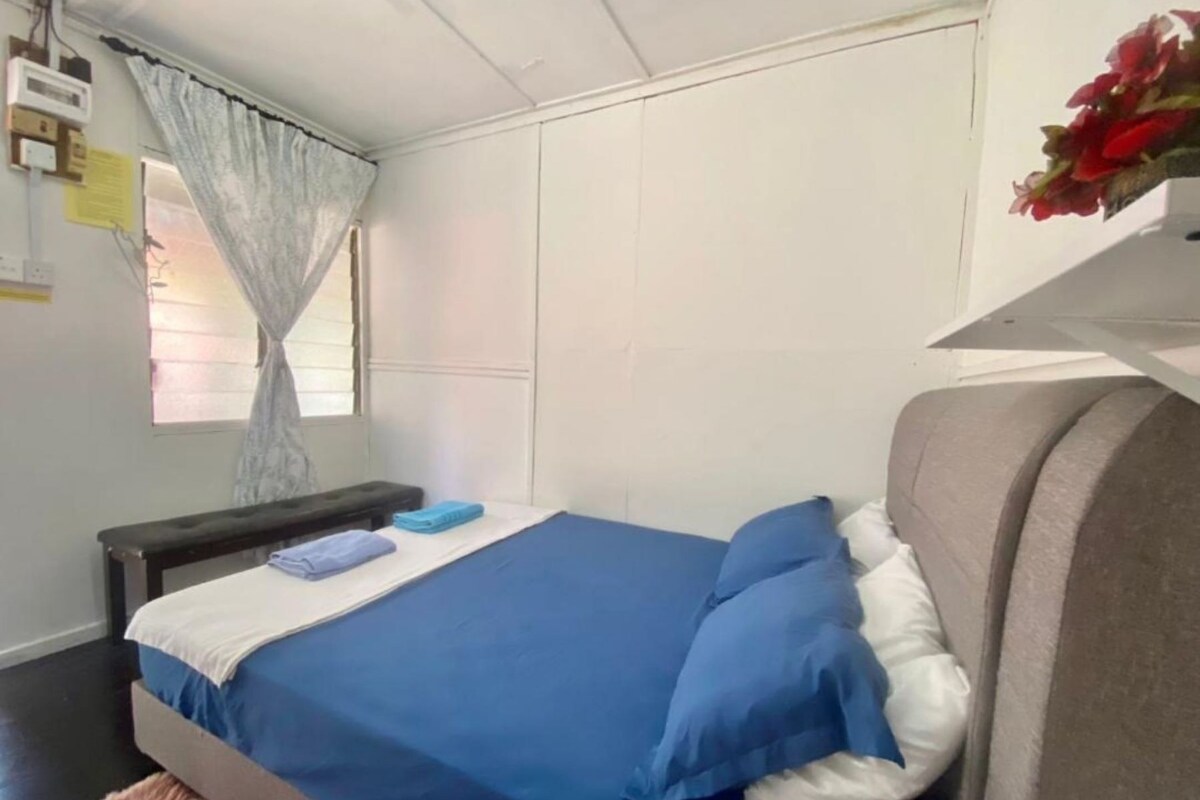 Una cama individual con ropa de cama azul, una mesita de noche, un banco y una ventana con cortinas.
