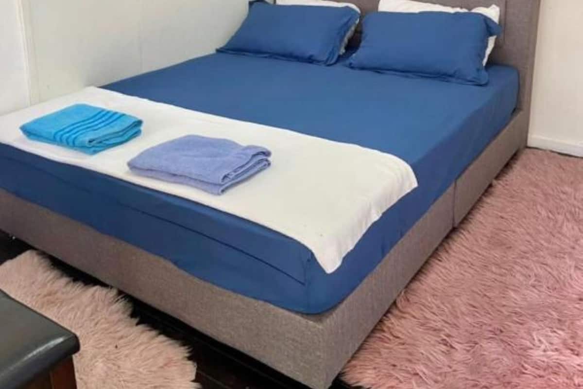Una cama individual con cabecera gris y ropa de cama azul, un felpudo rosa y una pequeña mesa con una silla.