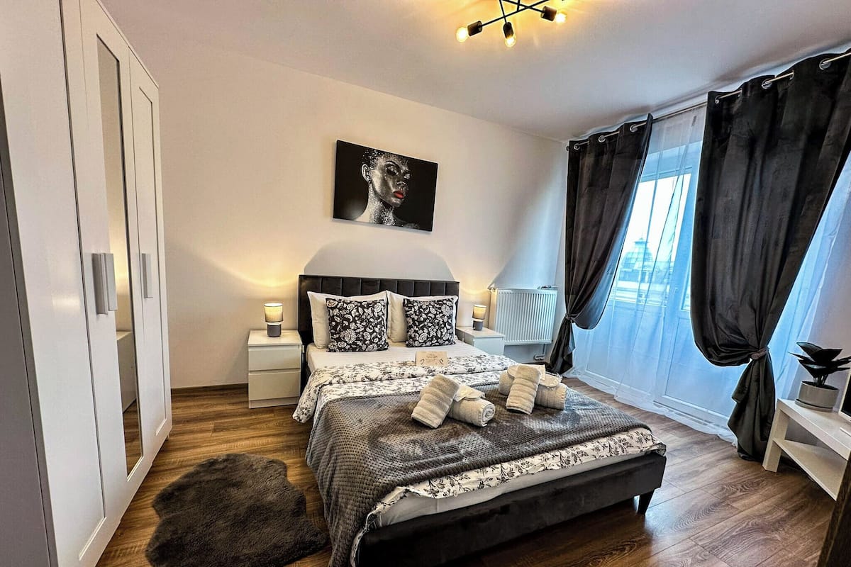 Ein modernes Schlafzimmer mit einem großen Bett, einem Kopfteil, einem Nachttisch mit Lampe, einem Beistelltisch, einem schwarzen, flauschigen Teppich, einem Fenster mit Vorhängen und einem Wandbild.