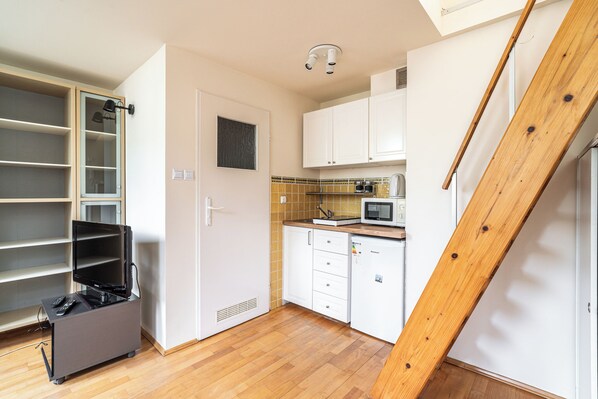 A compact kitchen with white cabinets, a microwave, and a refrigerator. There's a wooden staircase on the right and a black TV stand on the left. - RentPlanet - Apartament Garbarska (Kraków)