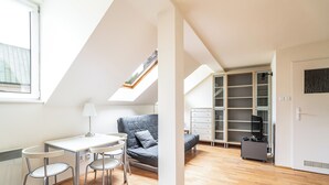 A modern, well-lit room with a skylight, a gray sofa, a dining table with chairs, and built-in shelving. - RentPlanet - Apartament Garbarska (Kraków)