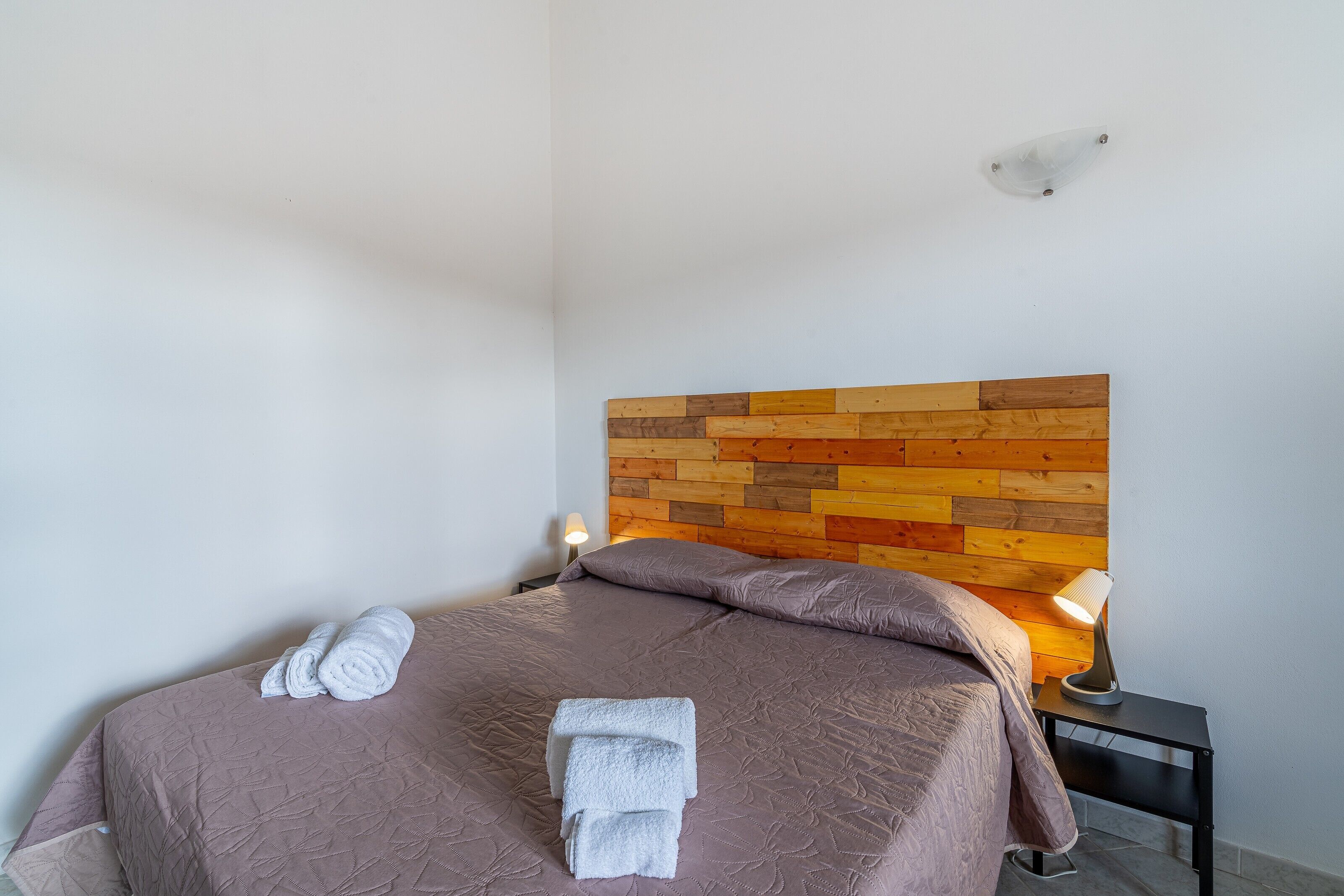 A neatly made bed with a headboard, a bedside lamp, and rolled towels.