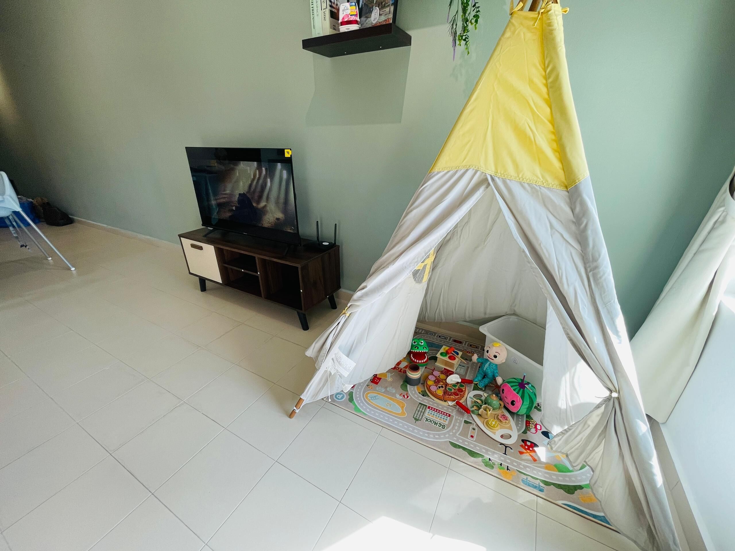 A room with a play tent, a television on a wooden stand, and a shelf with books and a plant.