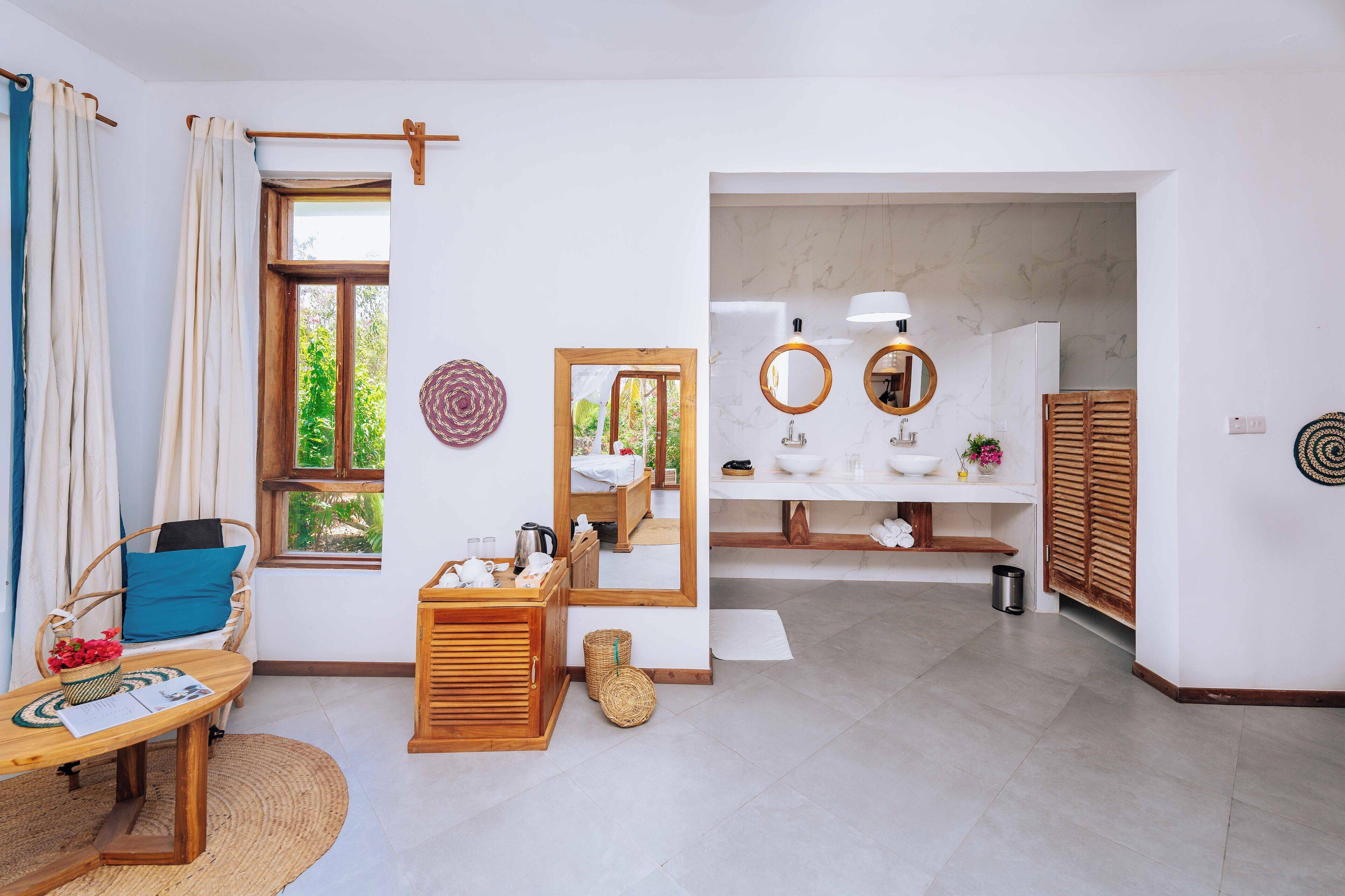 A modern room with a wooden dresser, a wicker chair, a small table with a basket of flowers, a mirror, and a view of the bathroom with a sink and mirror.