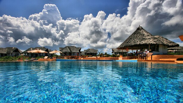 Una piscina con agua azul cristalina, rodeada de construcciones de techo de paja y un cielo despejado.