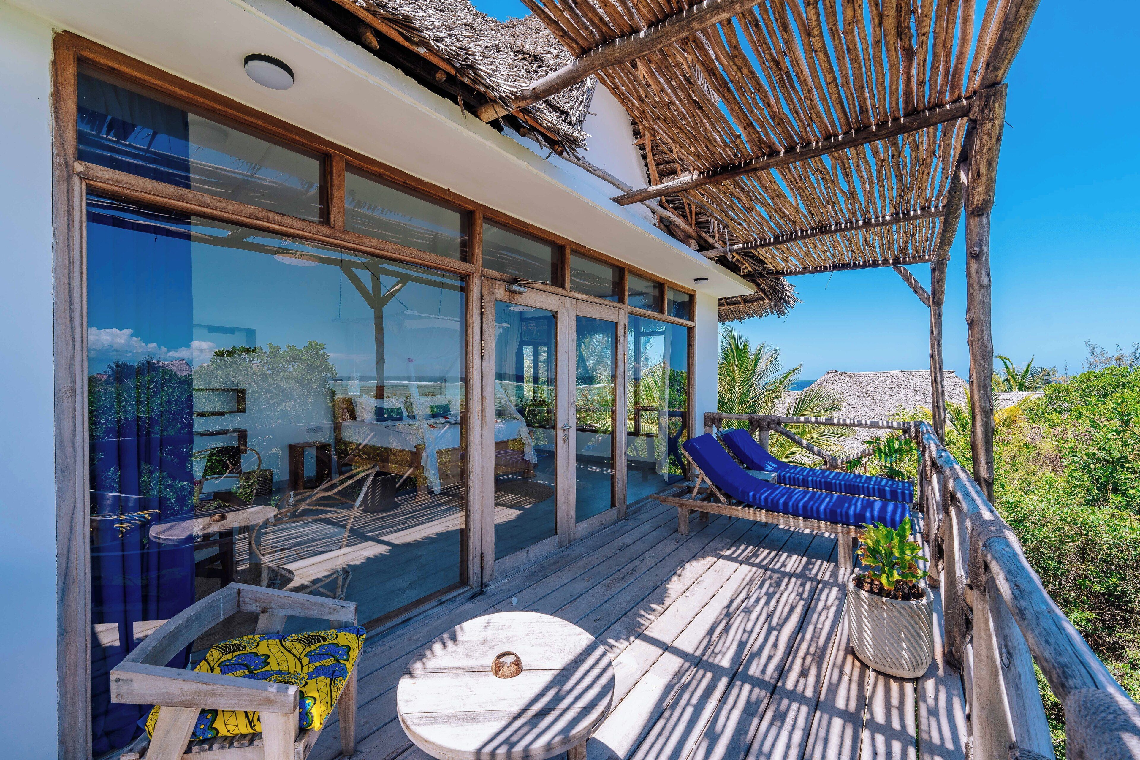Une terrasse en bois surmontée d’un toit de chaume, de grandes fenêtres en verre et un espace salon avec des coussins bleus.