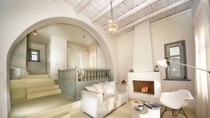 A spacious living area with a staircase leading to an upper floor, a white sofa, a white chair, a small table with books and a cup, and a fireplace. - White Village (Rhodes)