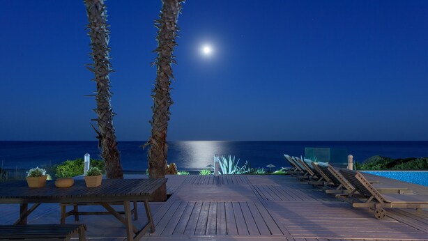 A wooden deck with lounge chairs and potted plants, overlooking a pool and the ocean under a night sky with a visible moon. - White Village (Rhodes)