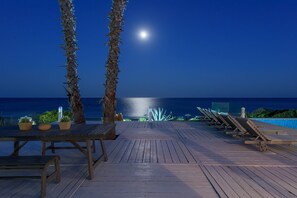 A wooden deck with lounge chairs and potted plants, overlooking a pool and the ocean under a night sky with a visible moon. - White Village (Rhodes)