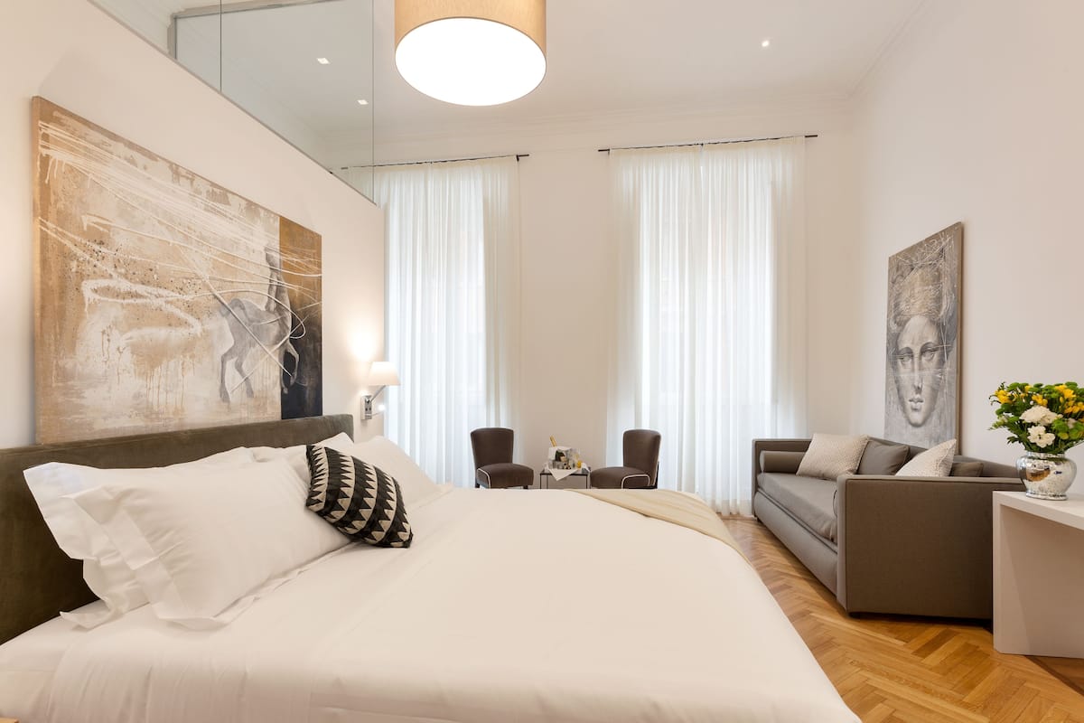 A modern hotel room with a large bed, a sofa, two armchairs, a small table, and artwork on the walls.
