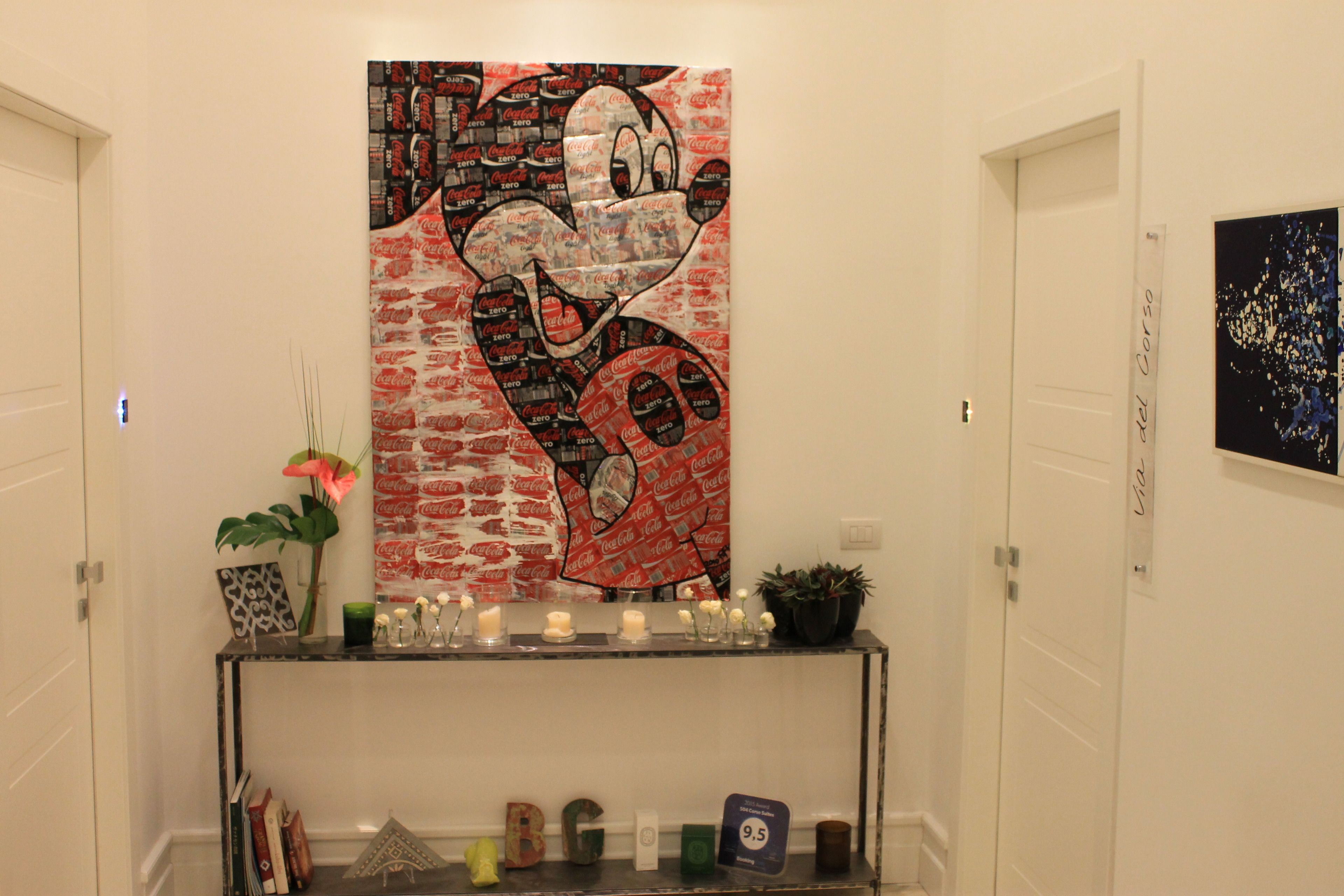 Un pasillo con una obra de arte temática de Coca-Cola en la pared, una consola con objetos decorativos y una planta en maceta.