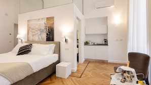 A modern hotel room with a bed, a small table with a bottle and glasses, a chair, and a kitchen area visible through an open door. - 504 Corso Suites (Rome)