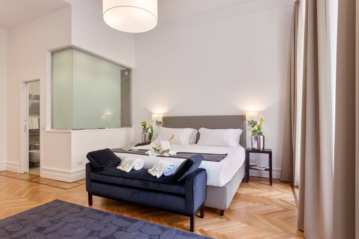 A modern hotel room with a large bed, a dark blue sofa, a small side table with a lamp, and a large window.