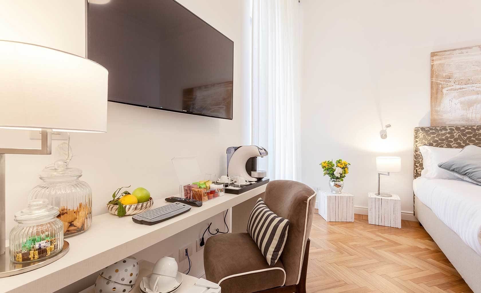 A modern hotel room with a desk, a chair, a bed, a TV, and a bedside table with a lamp.