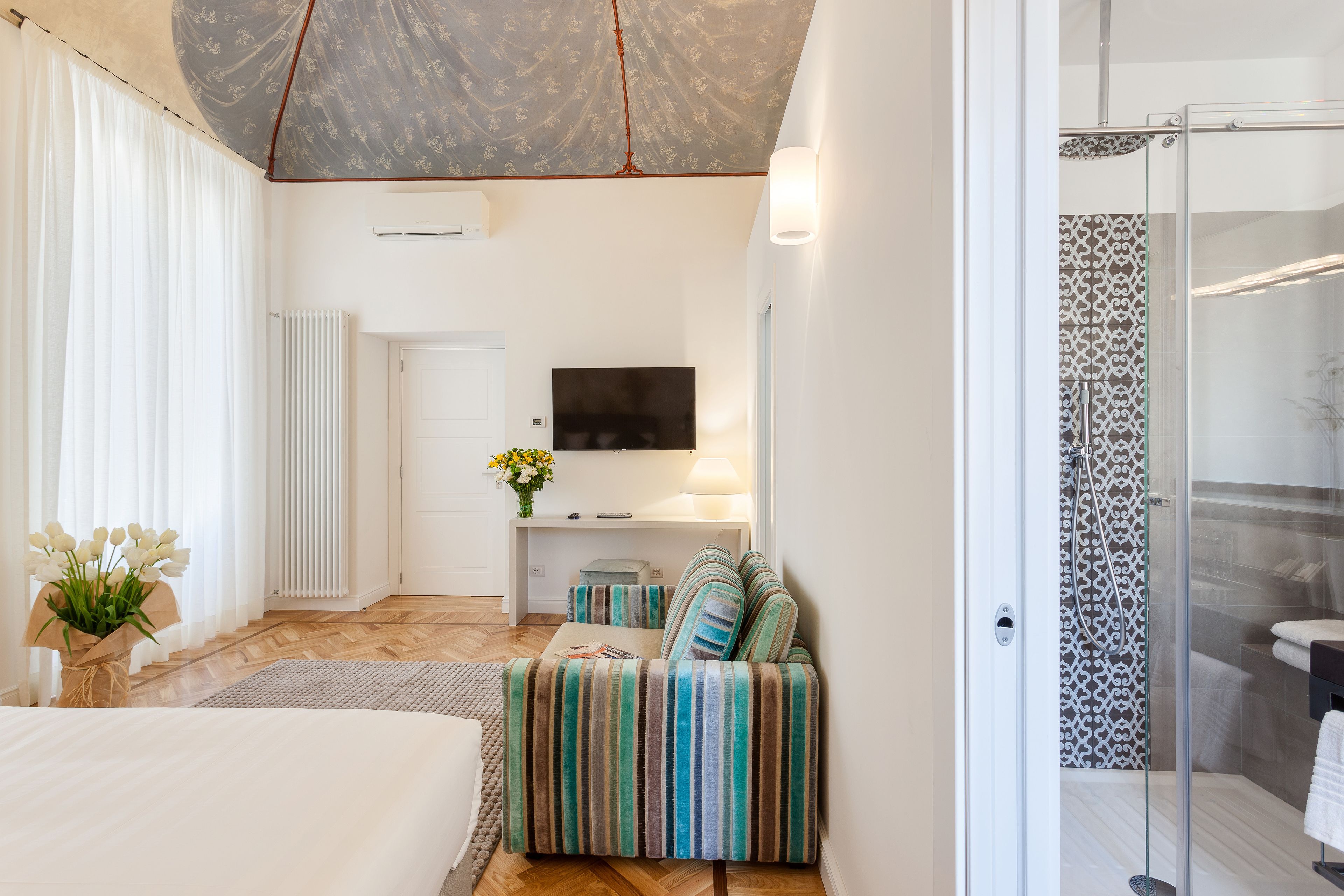 A modern hotel room with a bed, a sofa, a TV, a shower, and a small table with a vase of flowers.