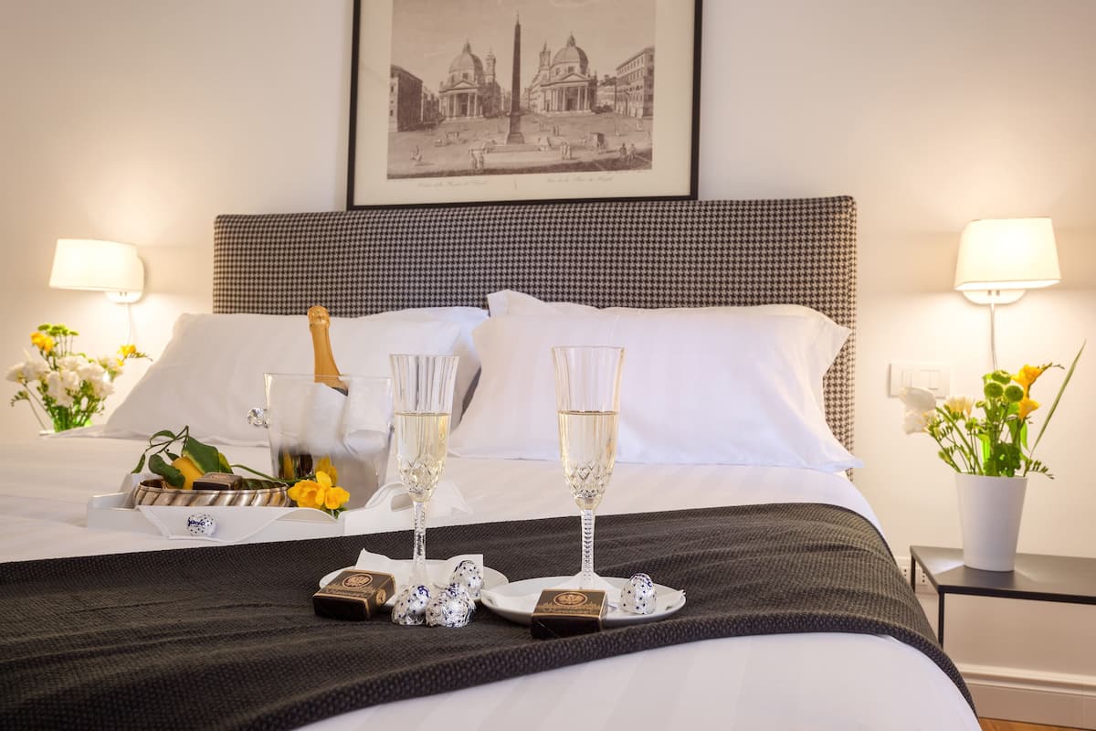 A neatly made bed with a checkered headboard, two bedside lamps, a framed cityscape picture, and a tray with champagne glasses, flowers, and chocolates on the bed.
