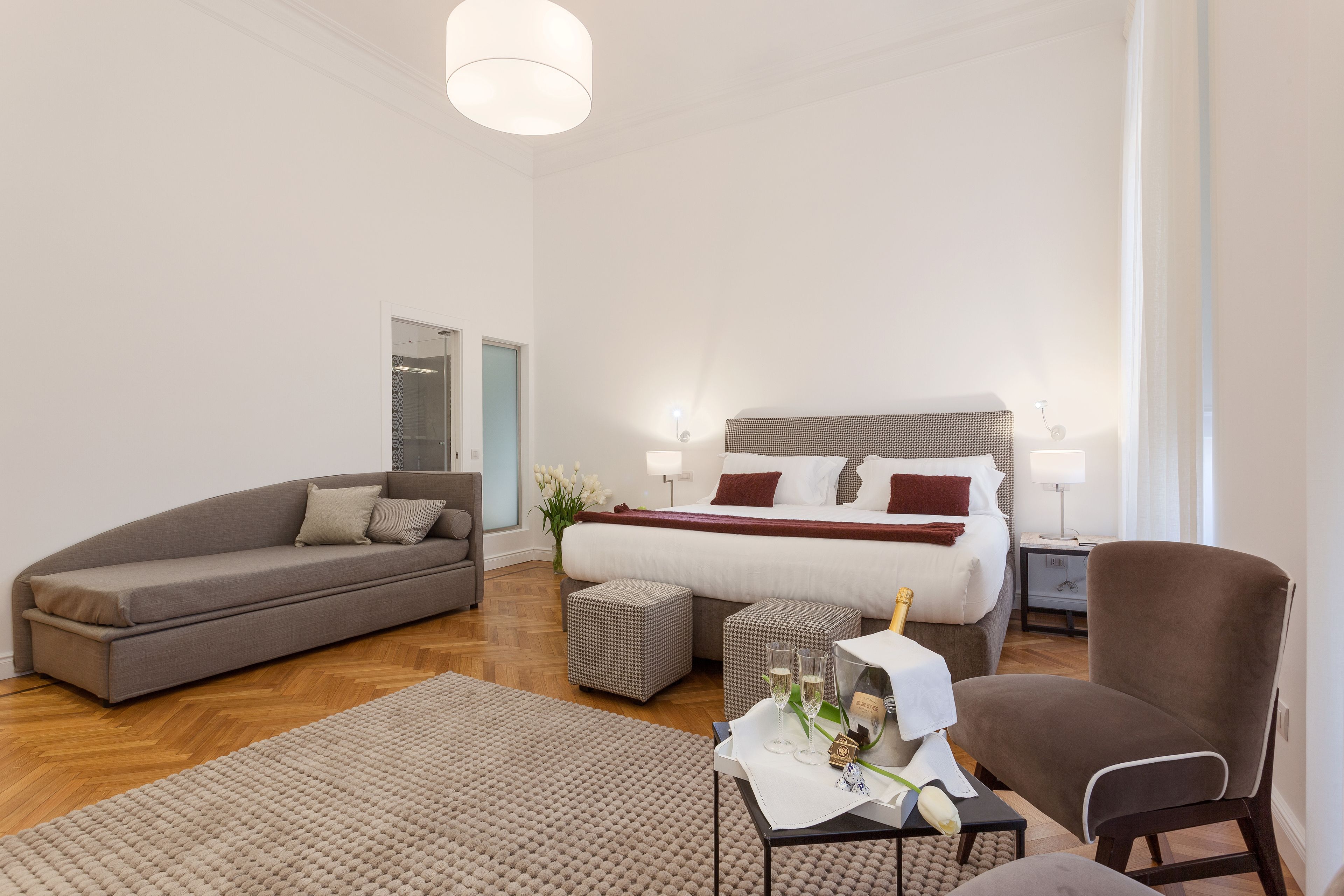 Ein modernes Hotelzimmer mit einem großen Bett, einem grauen Sofa, einem braunen Sessel und einem kleinen Tisch mit einer Vase und Gläsern.