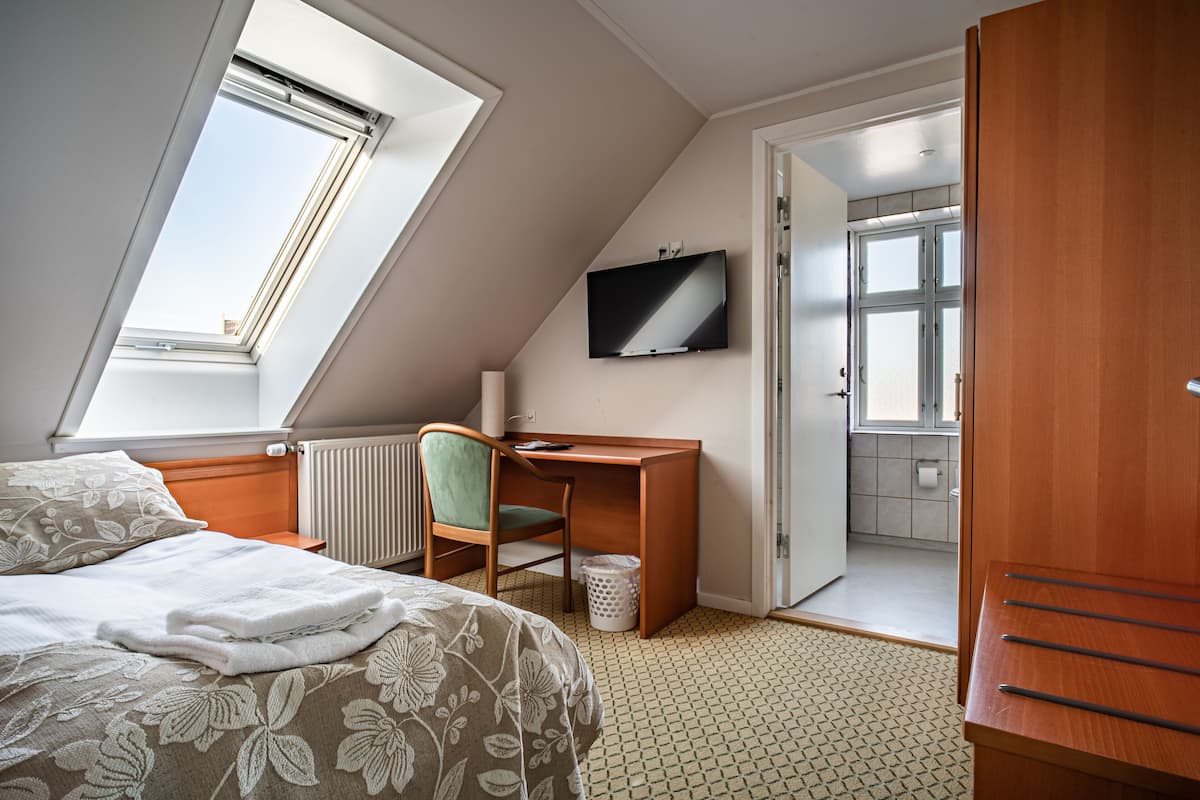A bedroom with a bed, a desk, a chair, a TV, and a bathroom visible through an open door.