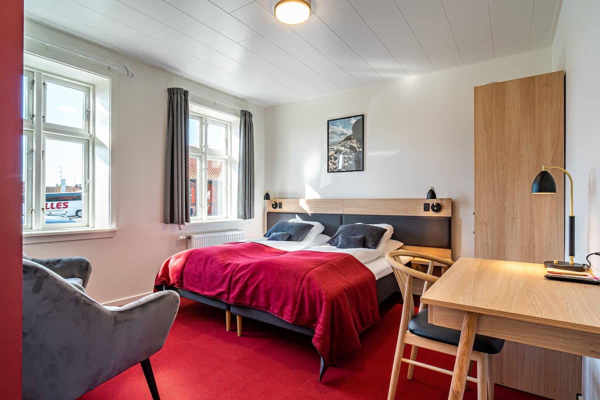 A bedroom with a bed, a desk, and a chair. There is a window with curtains, a radiator, and a picture on the wall.
