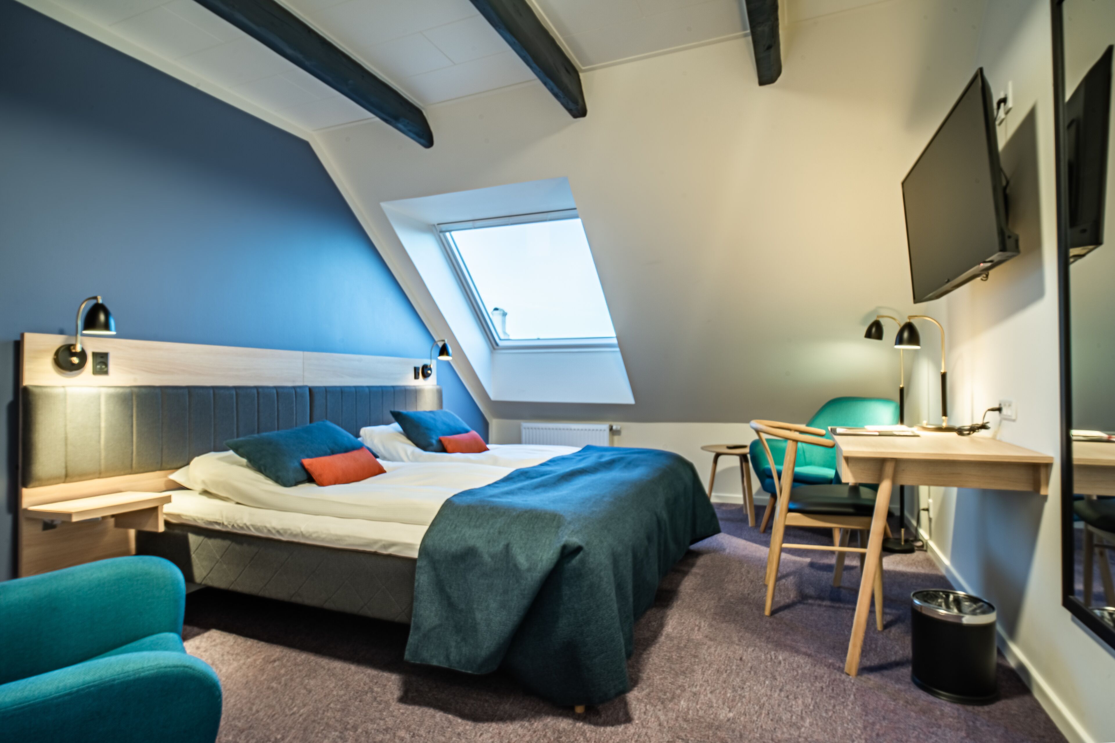 A bedroom with a sloped ceiling, a skylight, a bed with multiple pillows, a desk with a chair, and a TV mounted on the wall.