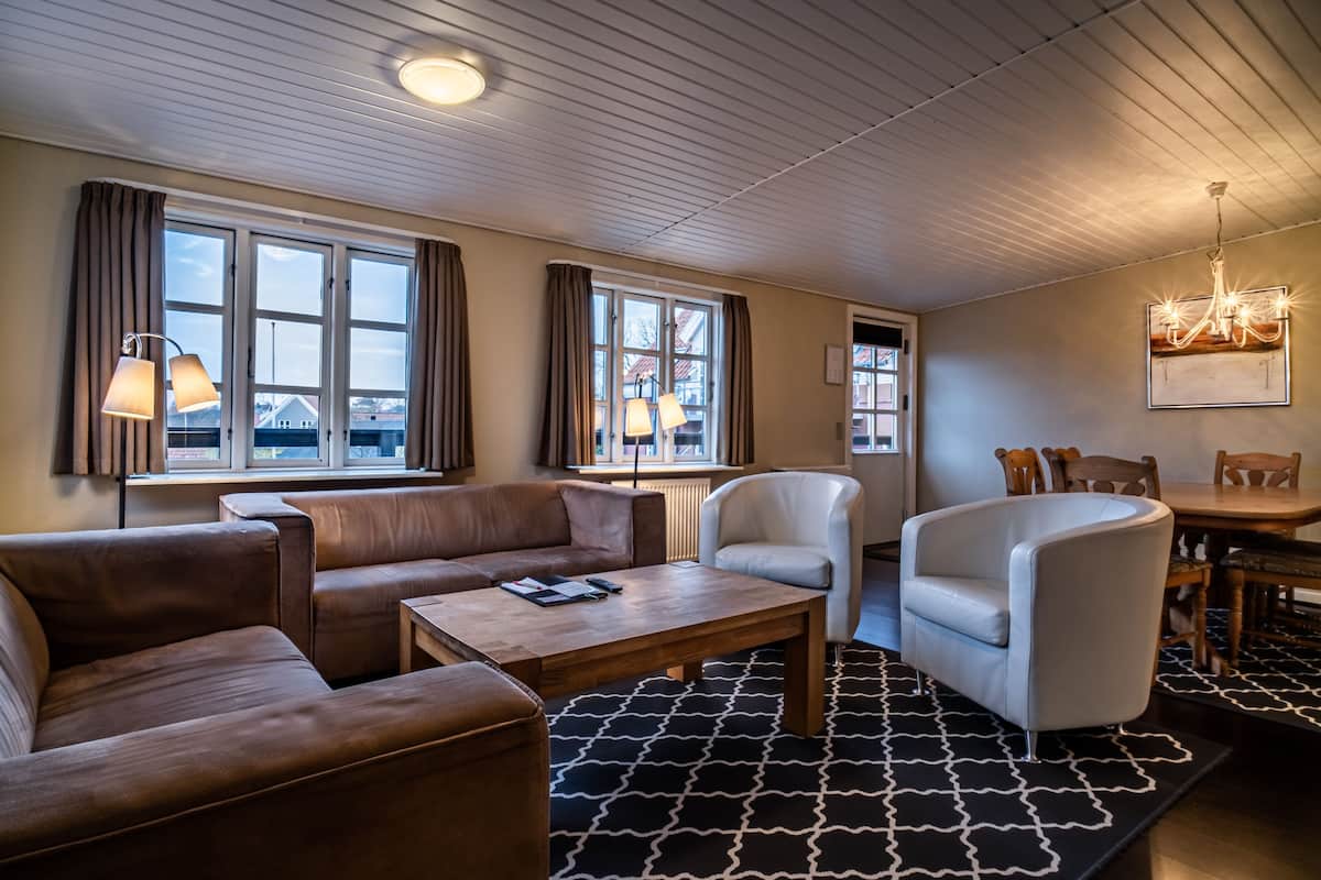 A modern living room with a sofa, armchair, and a coffee table. There are windows with curtains, a chandelier, and a dining area with a table and chairs.