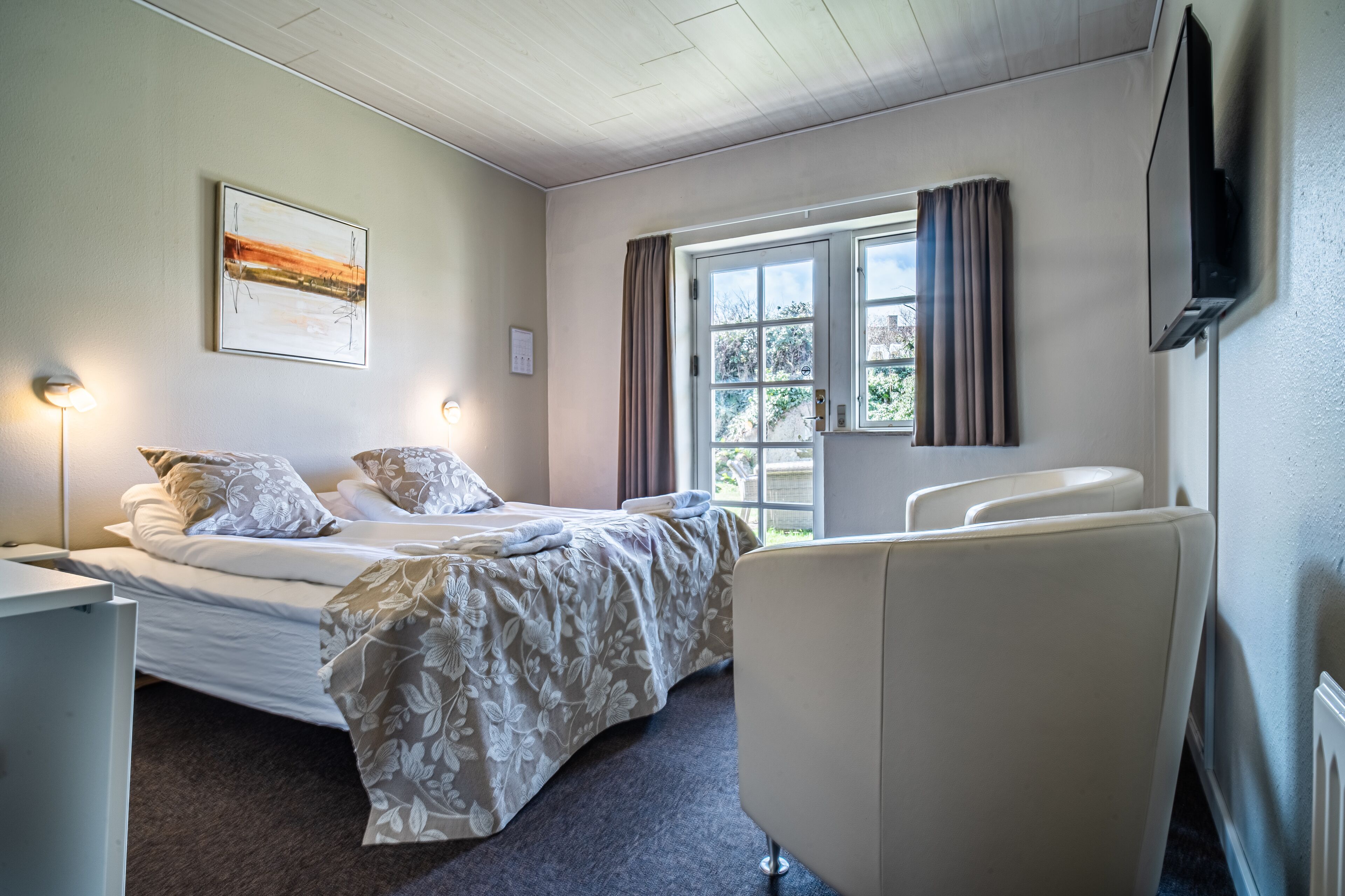 A hotel room with a bed, bedside table, lamp, window with curtains, and a TV mounted on the wall.