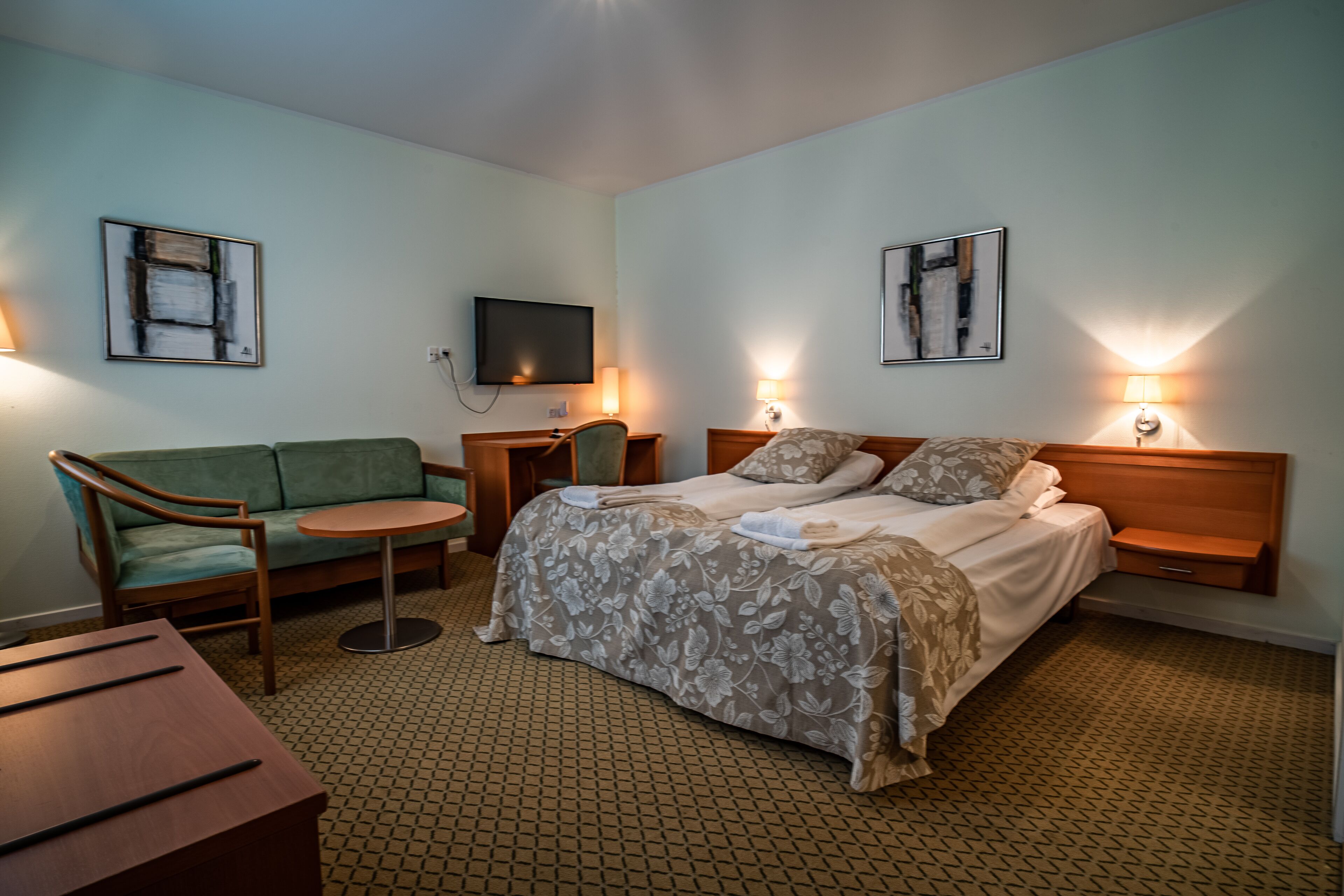 A hotel room with a bed, a sofa, a small table, and a TV on the wall.