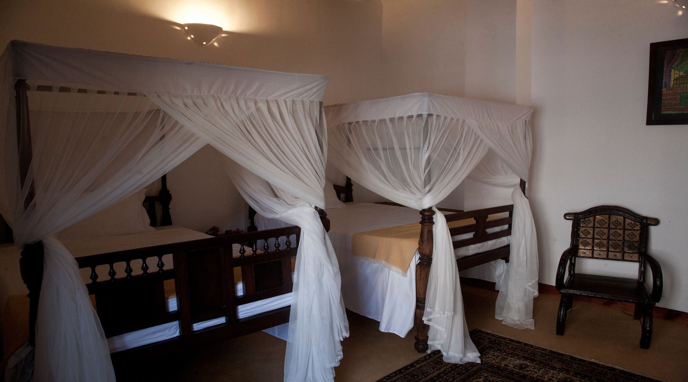 Een slaapkamer met twee houten bedden, elk voorzien van een hemelbed. Op de vloer staat een stoel en een vloerkleed.