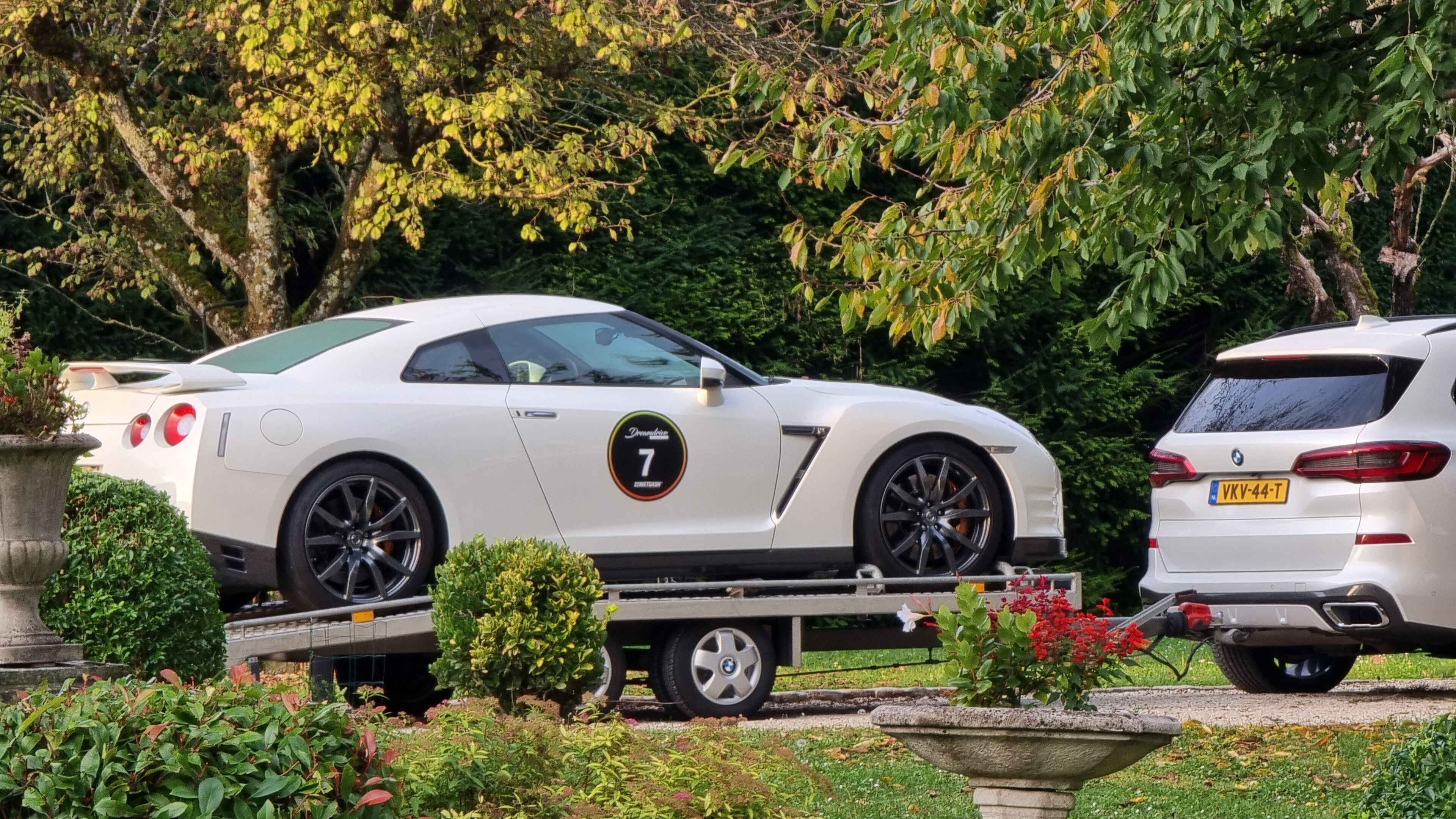 A white sports car on a trailer in a garden with greenery and a white car parked nearby.