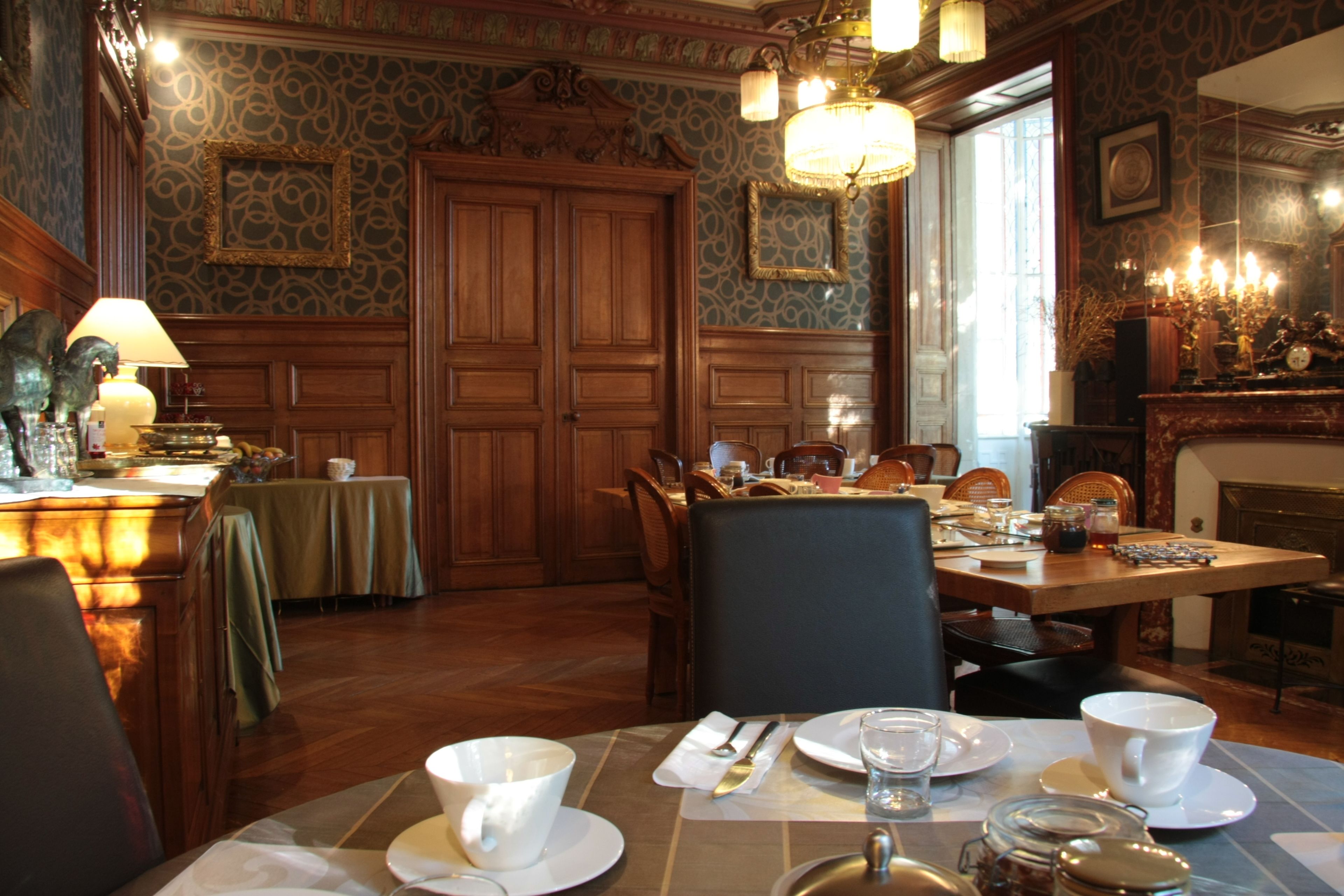 A dining area with a wooden table set for four, surrounded by chairs. There is a chandelier above the table, and a fireplace is visible in the background.