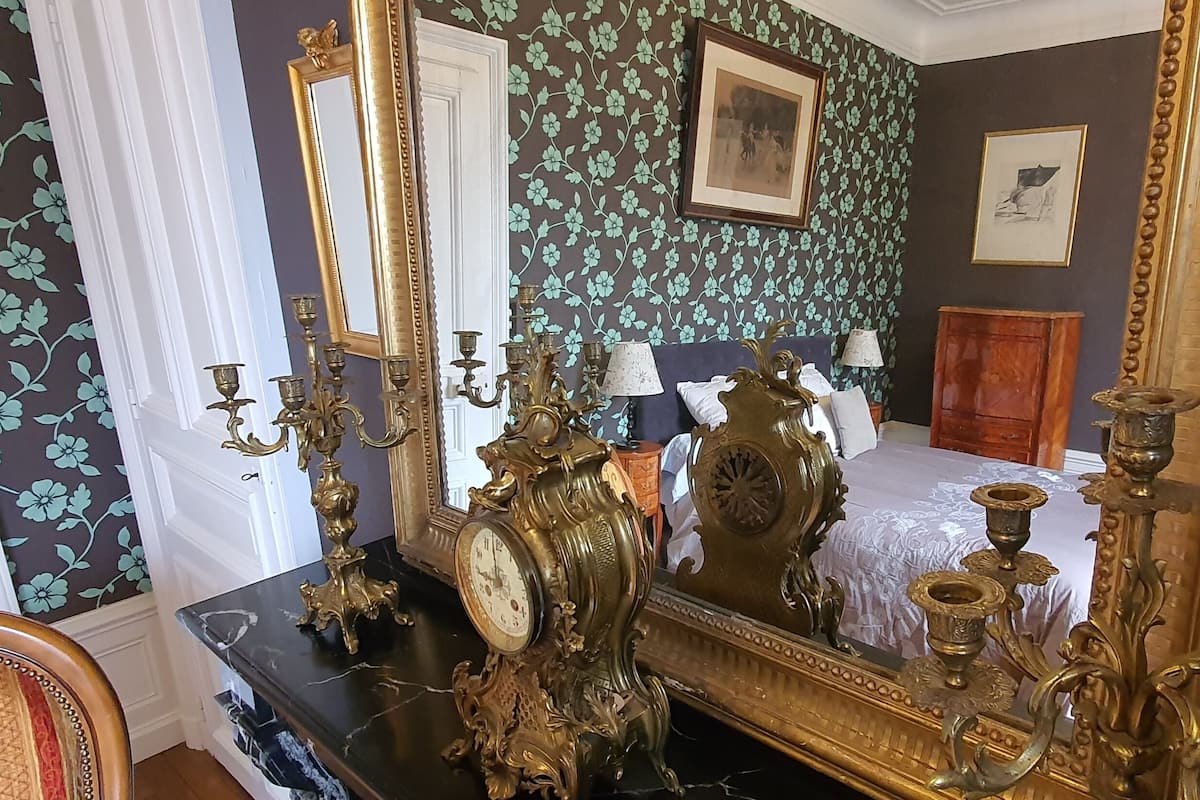 Um quarto com um espelho grande e ricamente decorado, uma cama com colcha branca e uma cômoda com espelho.