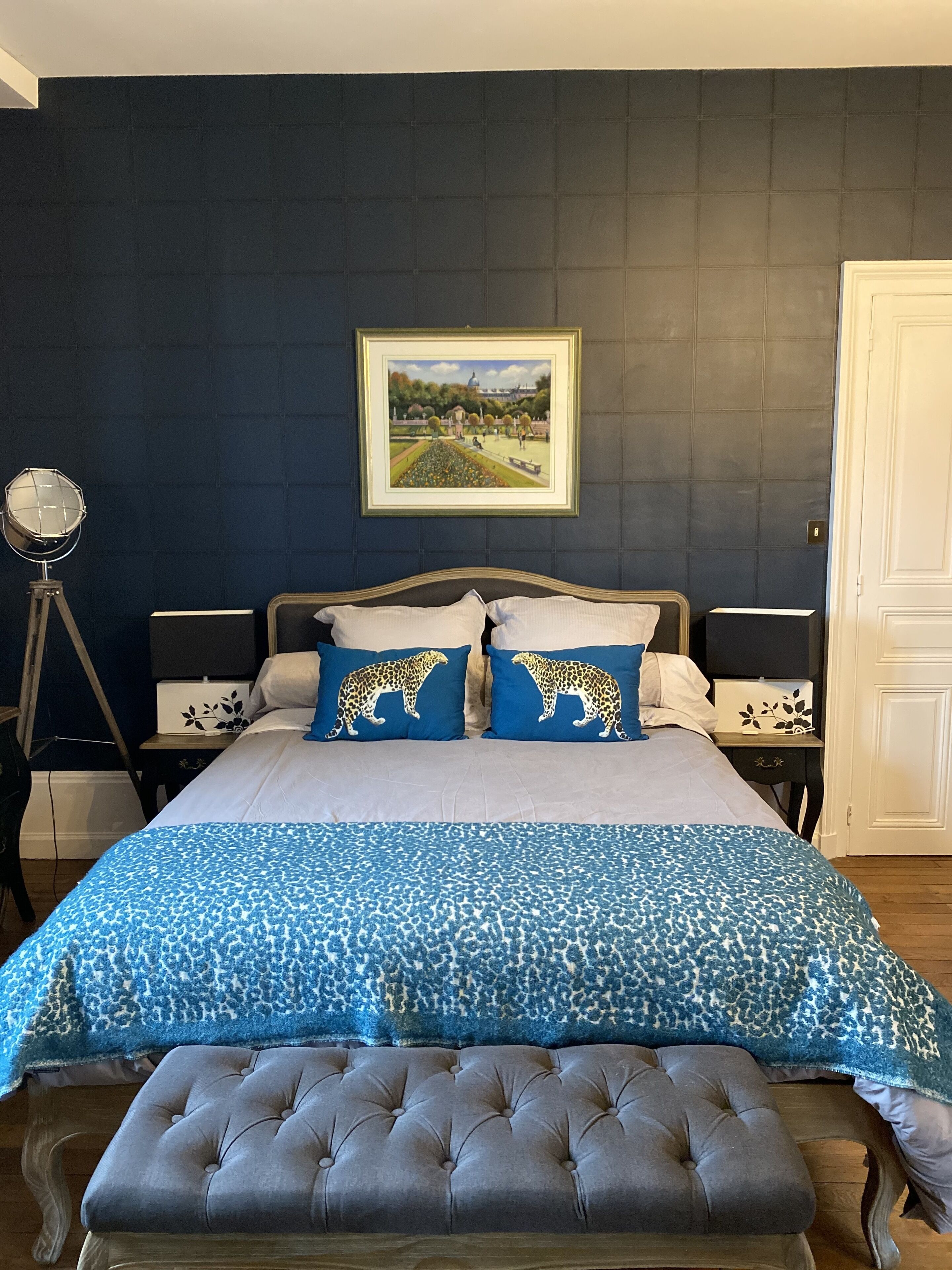A bedroom with a bed, two leopard-themed pillows, a blue patterned bedspread, a tufted bench, a framed painting on the wall, and a lamp on a tripod stand.
