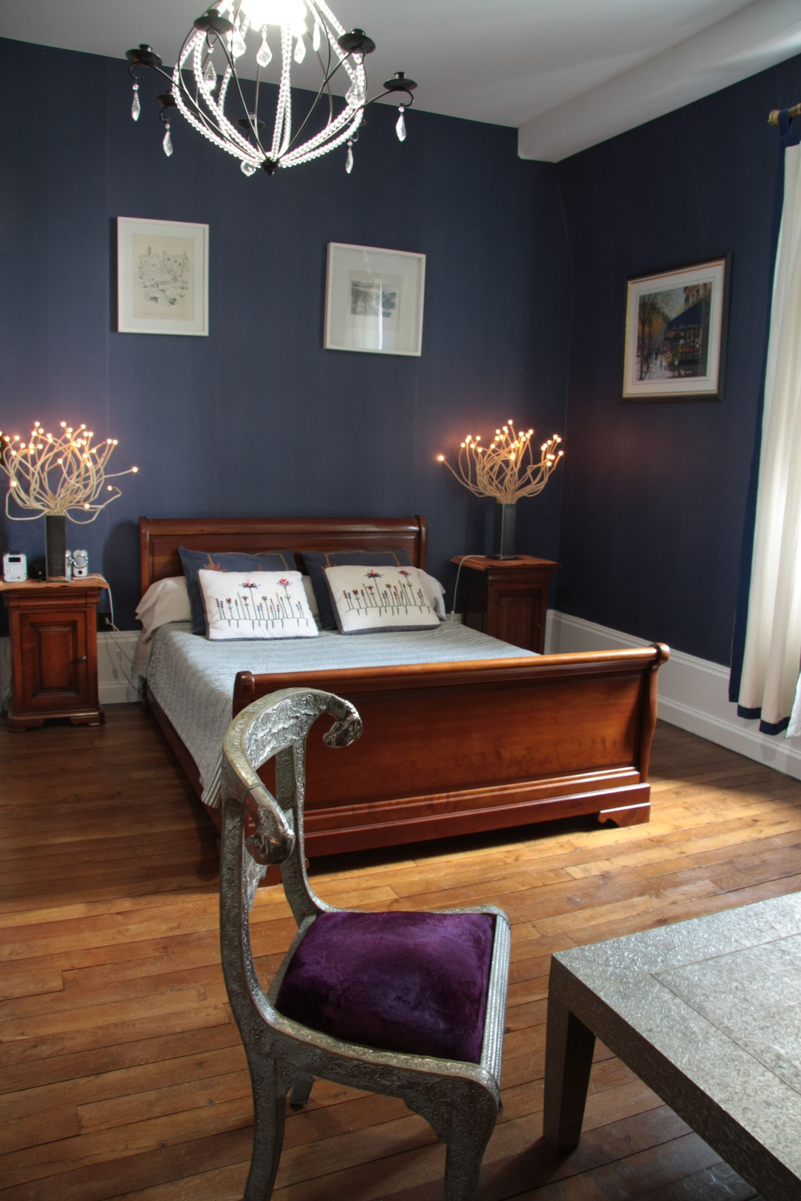 Ein Schlafzimmer mit einem großen Bett, einem hölzernen Kopfteil, zwei Nachttischen, einer Chandelieber und einem Stuhl mit violettem Kissen.