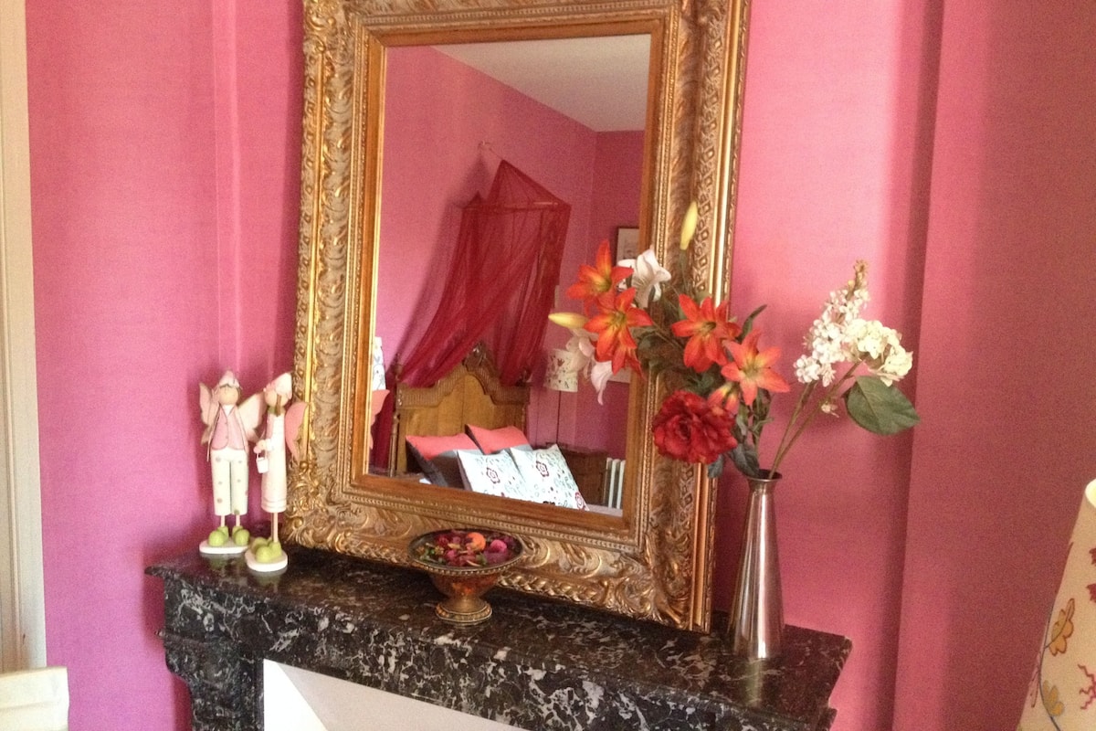 Quarto com um espelho grande e ornamentado sobre uma bancada de mármore, um vaso com flores e uma pequena estatueta.